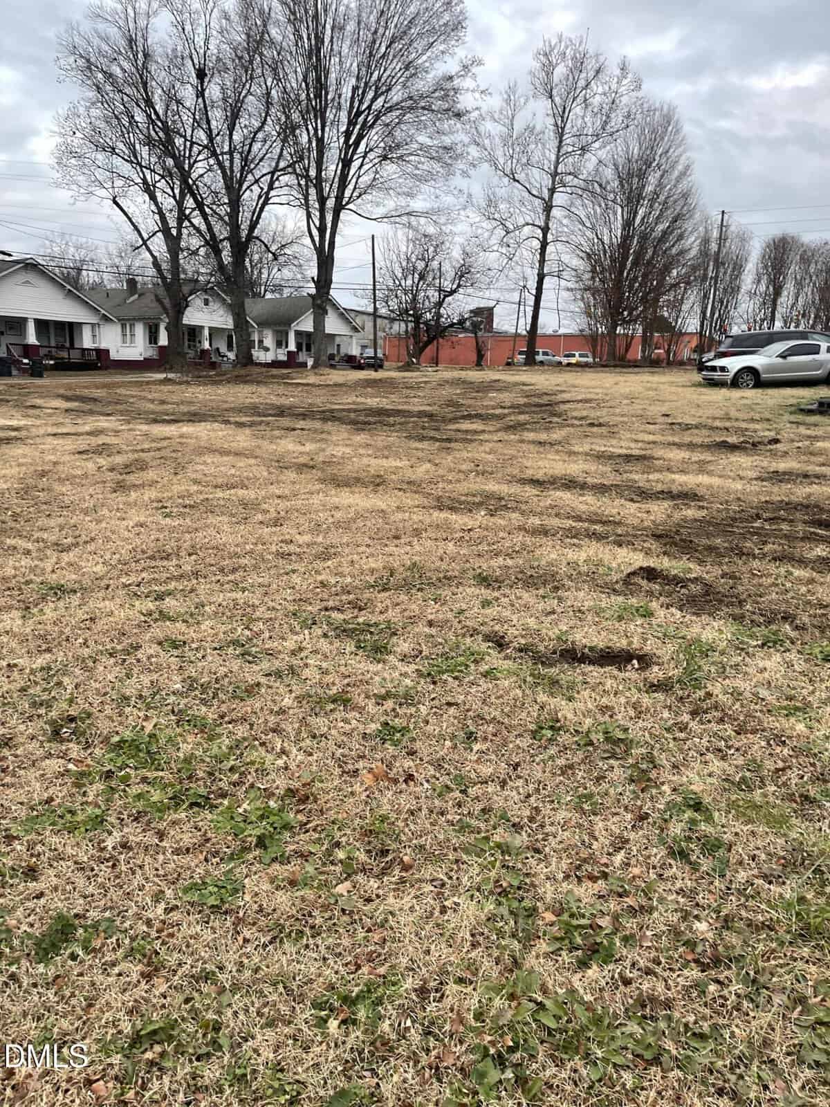 A vacant grassy lot with patches of brown grass, bordered by leafless trees. Houses and a few parked cars are visible in the background under an overcast sky.