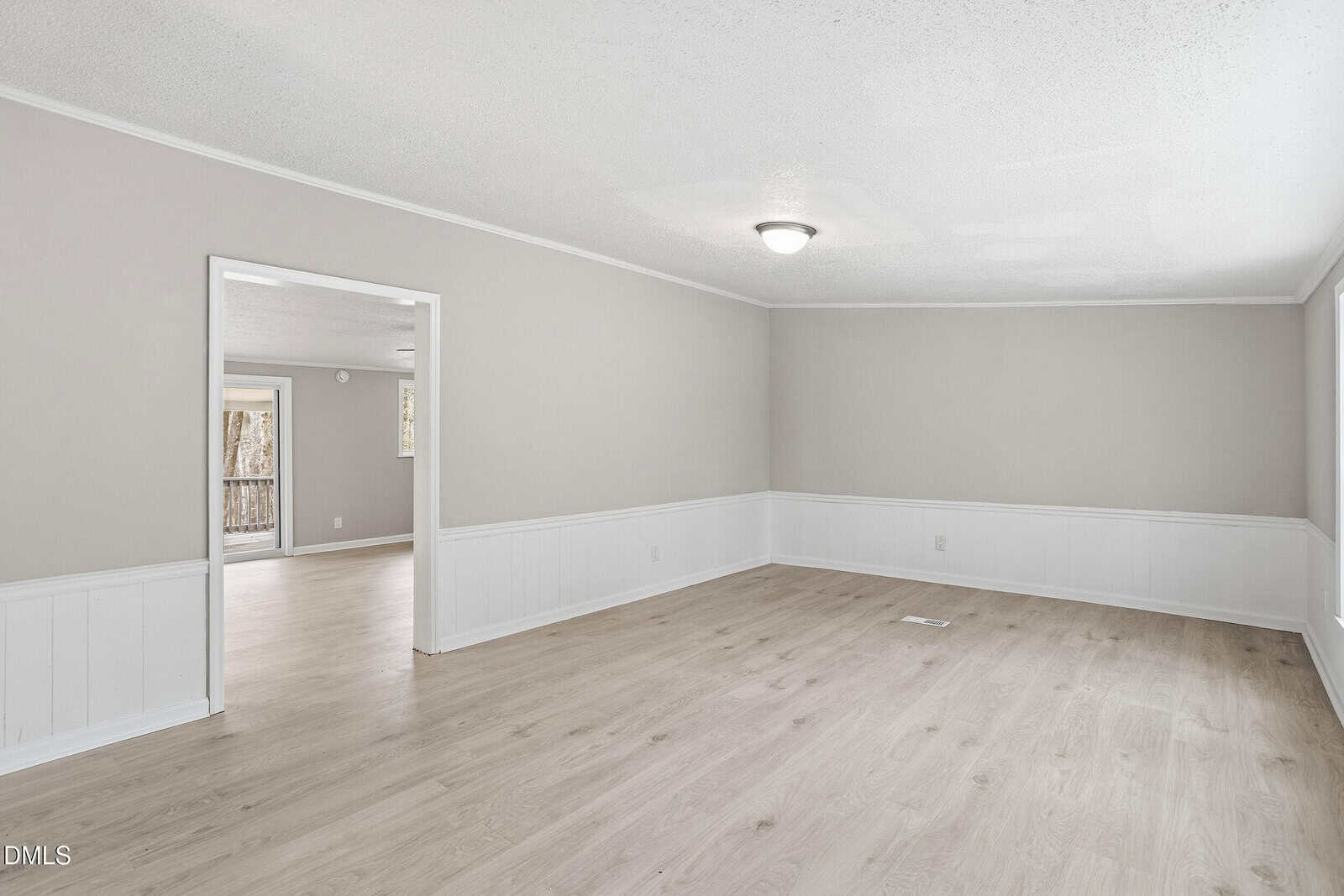 An empty room with light wood floors, beige upper walls, white wainscoting, and a ceiling light fixture. An open doorway leads to another room, and a window is visible in the adjacent space.