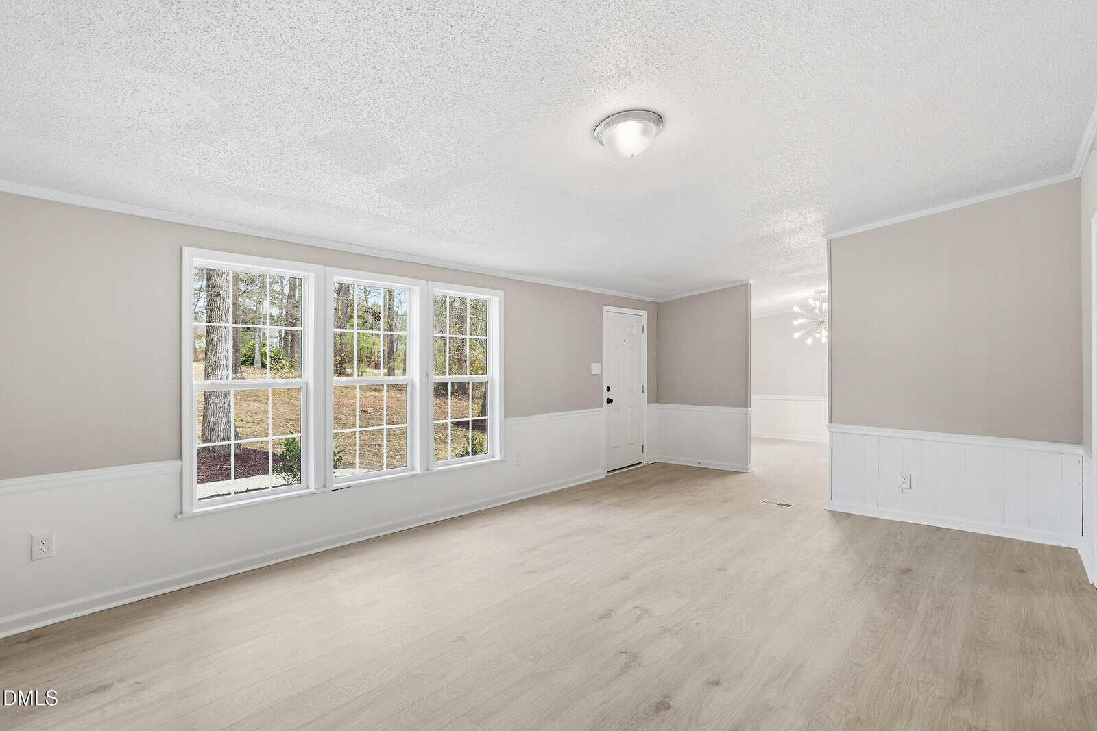 A bright, empty living room with light wood flooring, beige walls, white trim, three large windows, and a white front door. Sunlight fills the room, and a dining area is visible in the background.