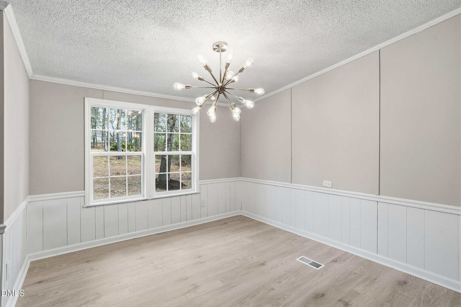 A bright, empty room with beige walls, white wainscoting, light wood flooring, a large window, and a modern chandelier on the ceiling. Sunlight streams in through the window.