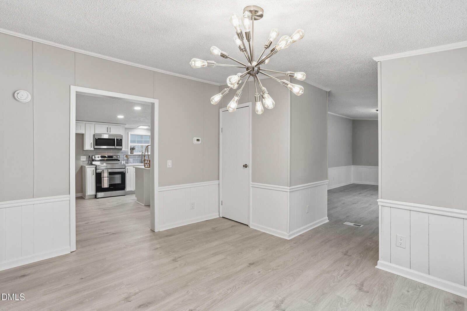 A modern dining area with light wood flooring, beige walls, and white wainscoting, features a sputnik-style chandelier. An open doorway leads to a kitchen with stainless steel appliances.