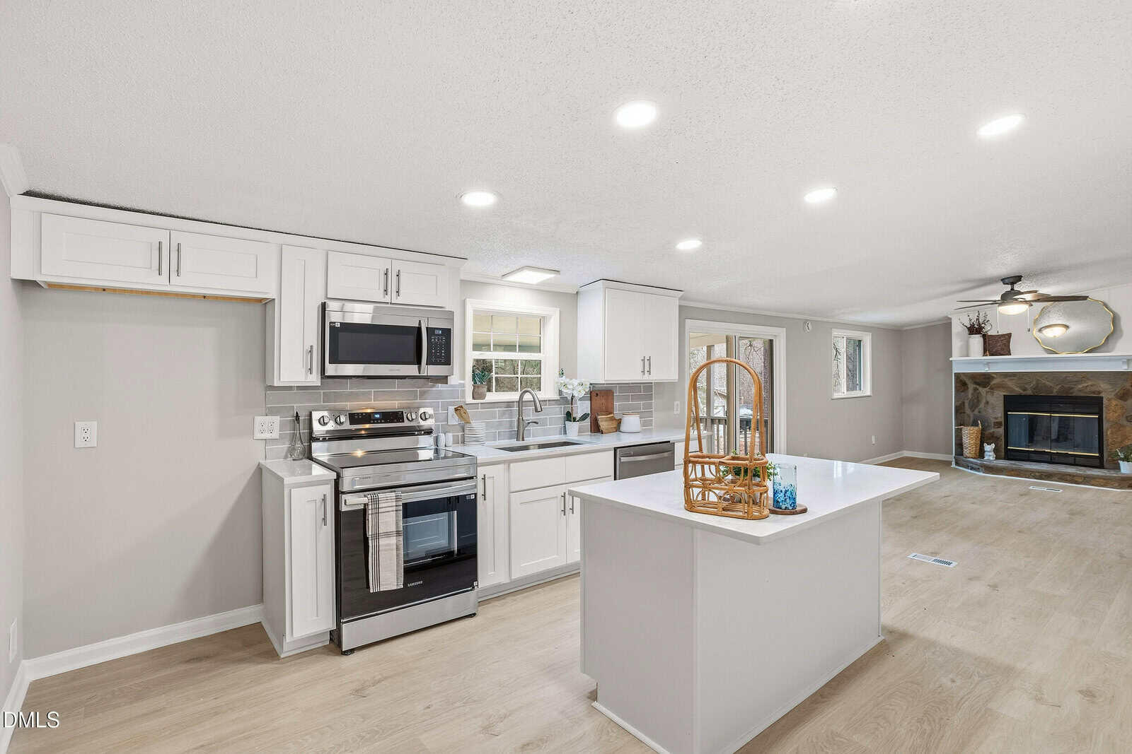 A modern kitchen with white cabinets, stainless steel appliances, an island with a basket tray, light wood flooring, and a view into a living area with a fireplace and ceiling fan.