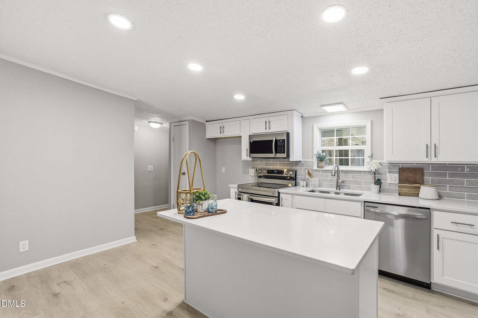 A modern kitchen with white cabinets, stainless steel appliances, a gray tile backsplash, light wood flooring, recessed lighting, and a white island with a tray holding decorative items. A window is above the sink.