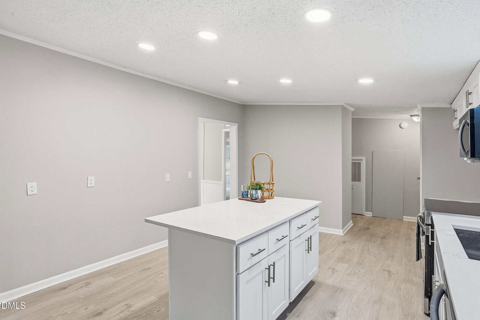 A modern kitchen with light wood floors, gray walls, white cabinets, and a white island in the center. The island has a few items on top, and recessed ceiling lights provide illumination.