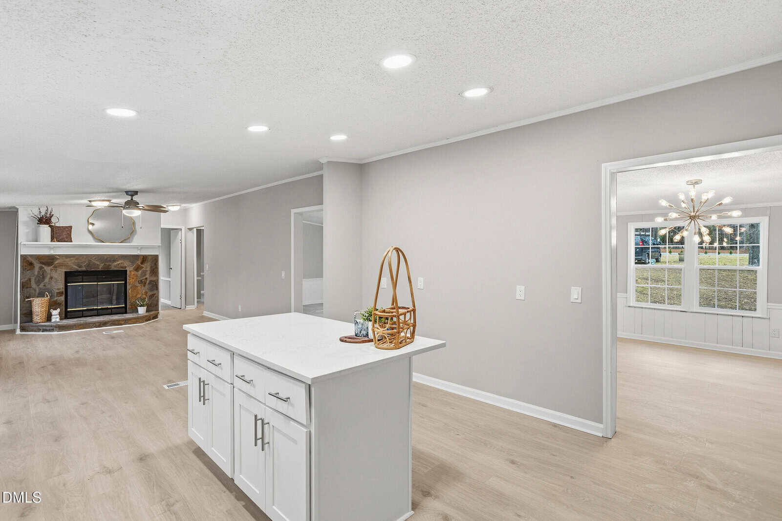 A modern, open-plan kitchen with a white island and cabinets, light wood flooring, and recessed lighting. A living area with a stone fireplace and a separate dining room are visible in the background.