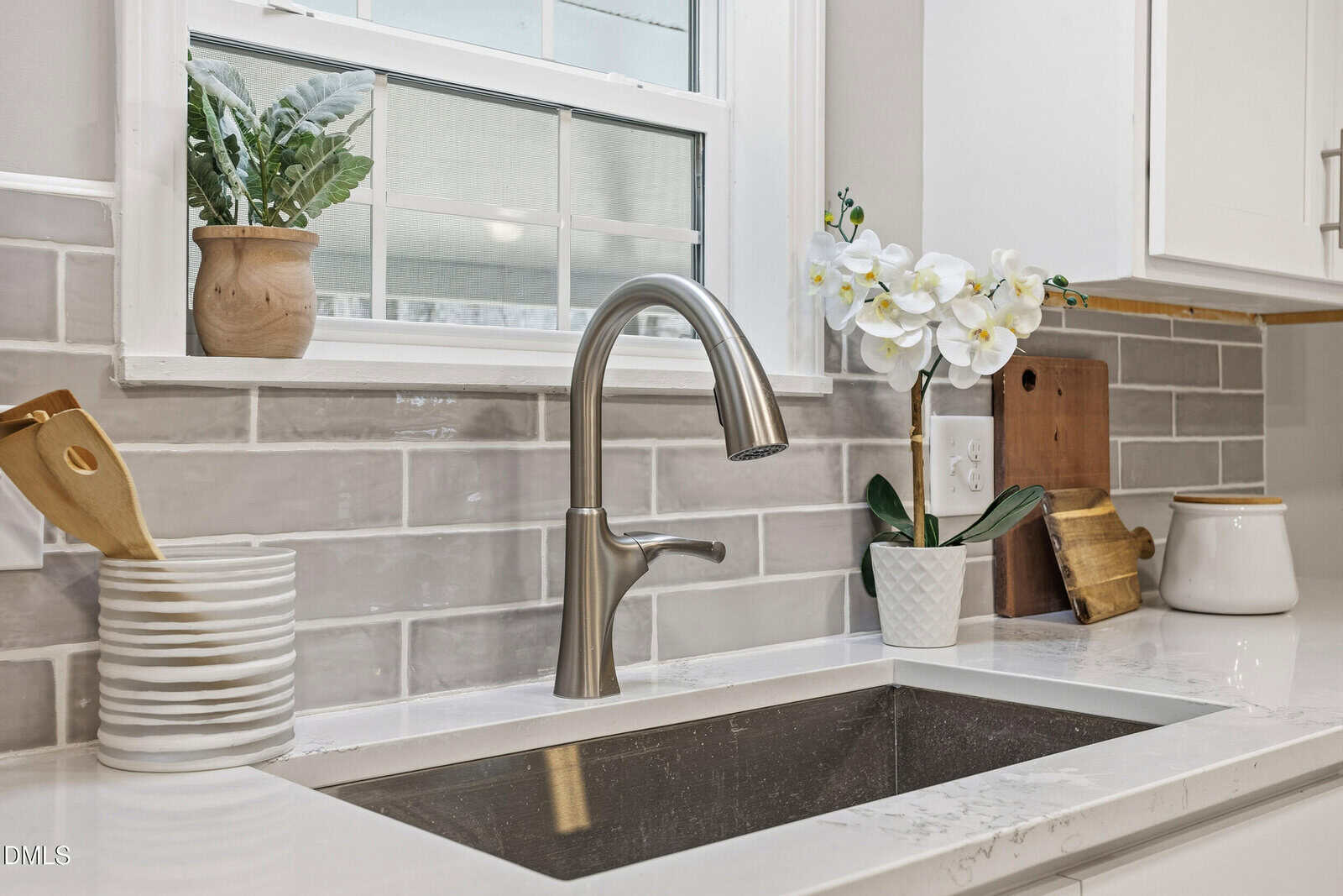 A modern kitchen sink with a stainless steel faucet, set in a white marble countertop. Gray tile backsplash, a potted plant, an orchid, stacked bowls, utensils, and a ceramic container are visible on the counter.