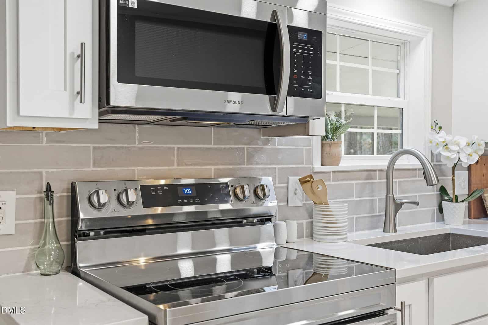 Modern kitchen with a stainless steel stove and microwave, white cabinets, tile backsplash, a sink with a faucet, stacked white plates, utensils in a holder, and a potted plant near a window.