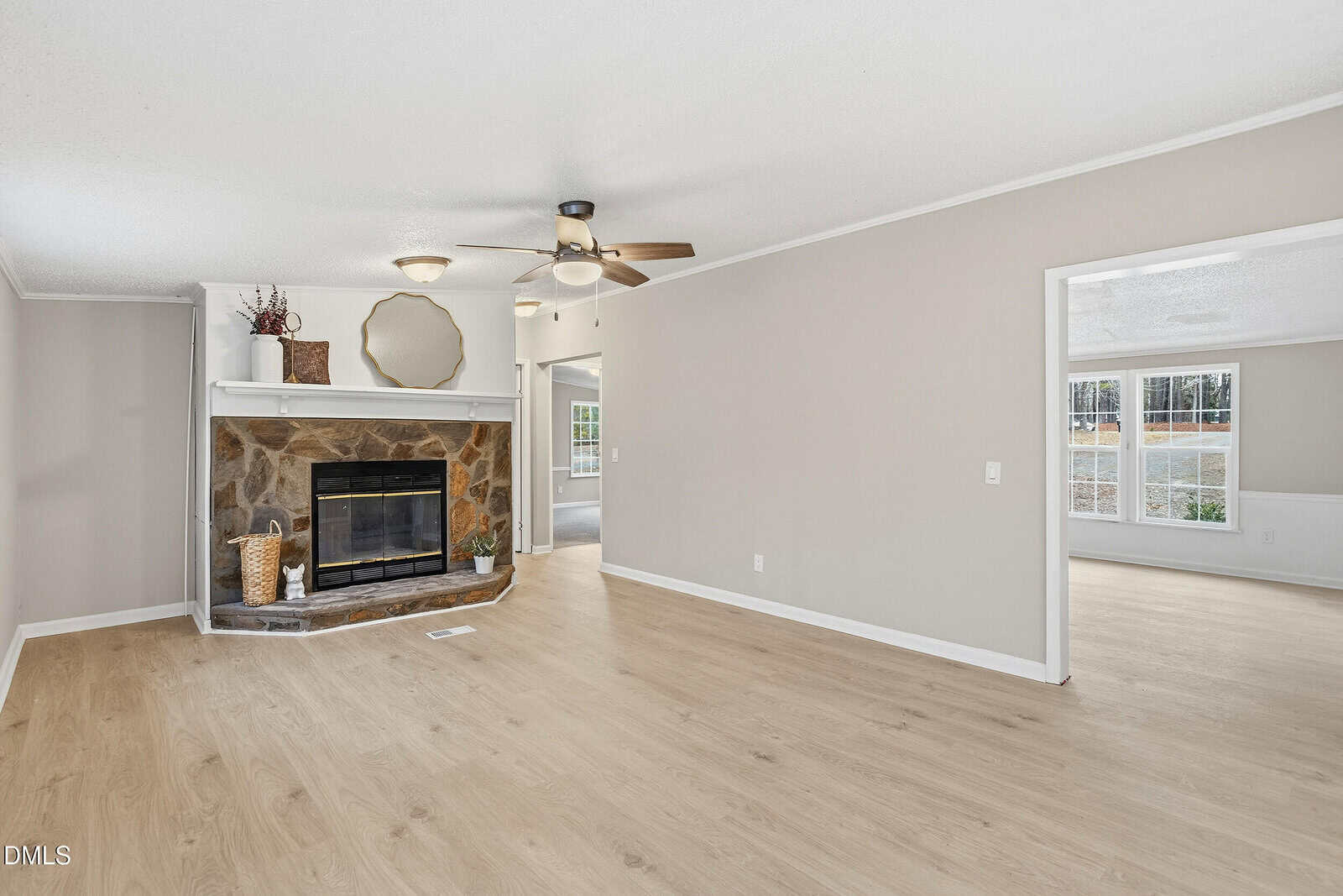 Bright living room with wood floors, stone fireplace, ceiling fan, and large windows. Neutral walls and an open doorway lead to another sunlit room. Minimalist decor with a round mirror and plants above the fireplace.