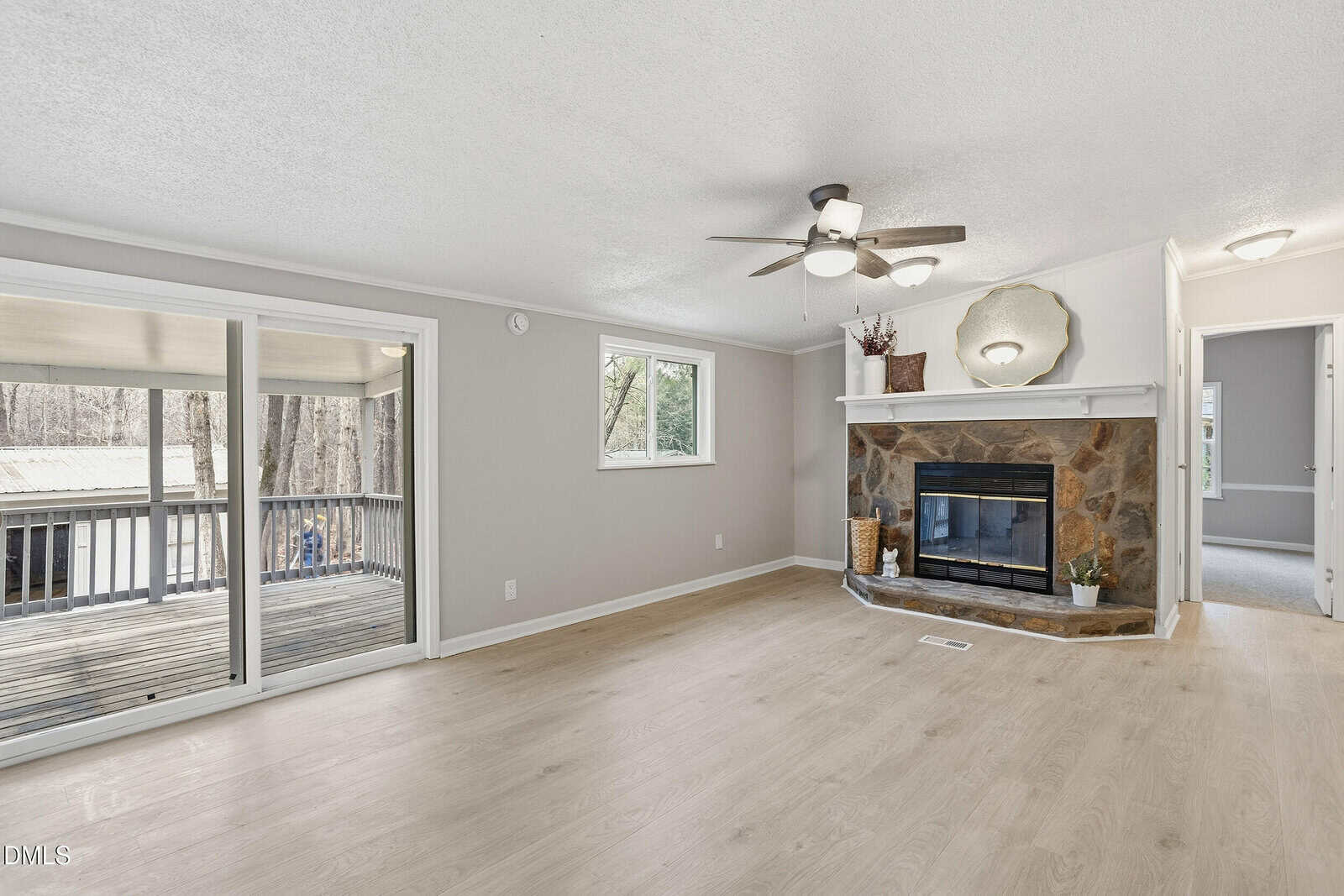 Bright living room with light wood flooring, stone fireplace, ceiling fan, and sliding glass doors opening to a covered deck with a wooded view. A hallway and small window are also visible.