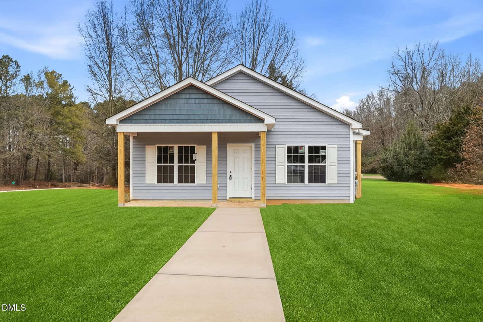 A small, single-story gray house with a gabled roof and wooden posts on the front porch, sits on a well-manicured green lawn, surrounded by trees under a bright blue sky.