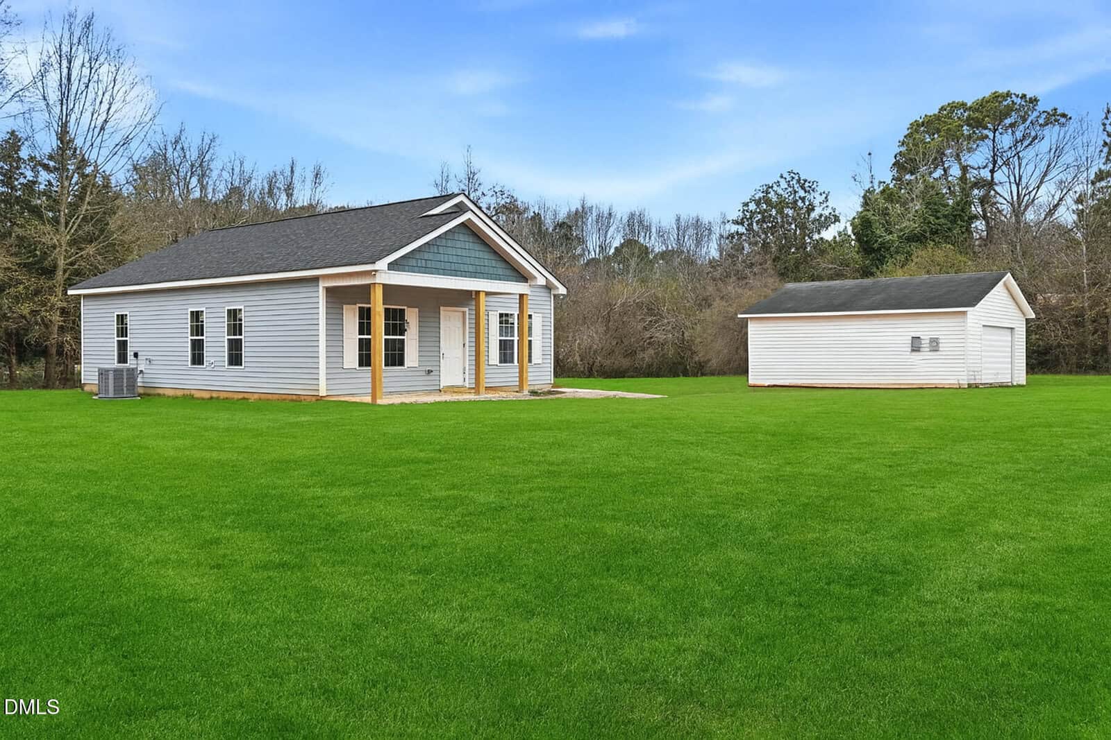 A small, single-story gray house with a covered front porch and white trim sits on a large, lush green lawn with a detached garage in the background and trees along the property line under a blue sky.