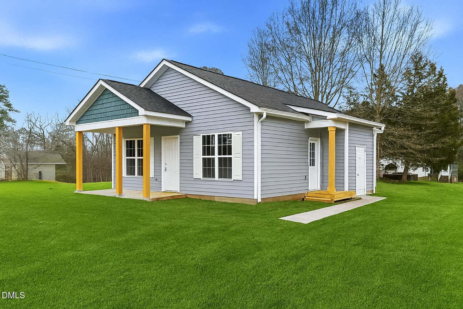 A small, single-story gray house with white trim and yellow columns, sitting on a large green lawn under a blue sky with trees in the background.
