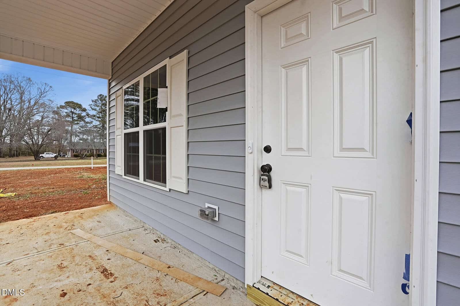 Close-up of a white front door and window with white shutters on a gray house exterior. A concrete porch and bare ground are visible, along with trees and houses in the background.