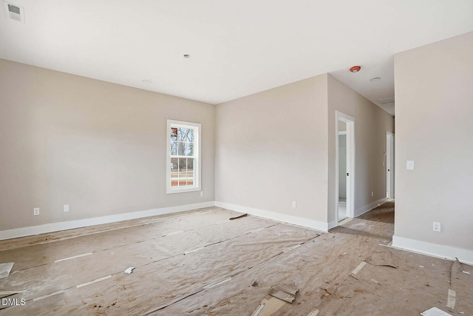 A spacious, empty room with beige walls and a window letting in natural light. The floor is covered with protective brown paper, and there are two open doorways leading to other rooms.