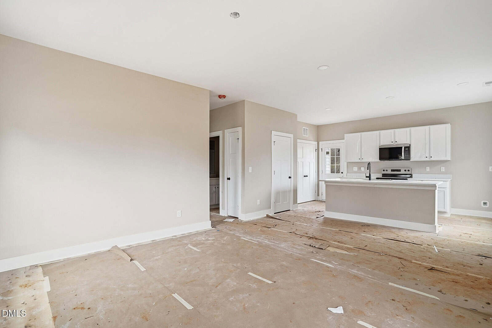 Unfurnished room with bare floors covered in protective paper, beige walls, and an open kitchen area with white cabinets, a stove, microwave, and a central island. Multiple doors and natural light from windows.