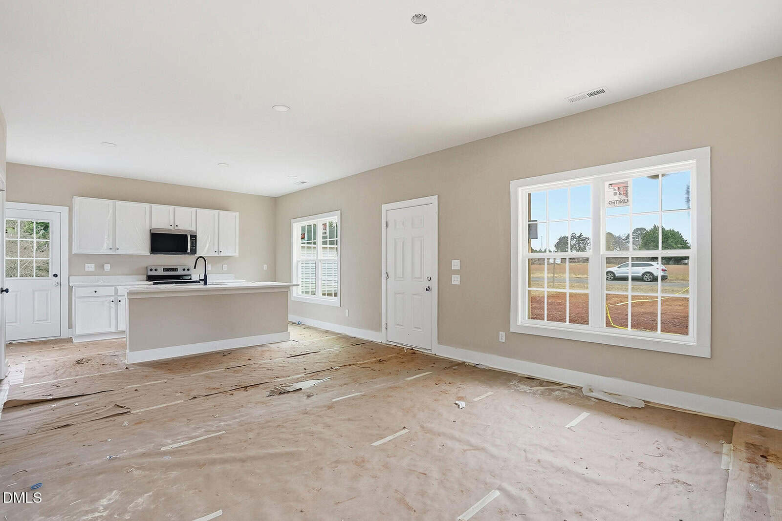 A bright, unfurnished kitchen and living area with white cabinets, stainless steel appliances, large windows, and tan walls. The floor is covered with protective paper, and a white exterior door leads outside.