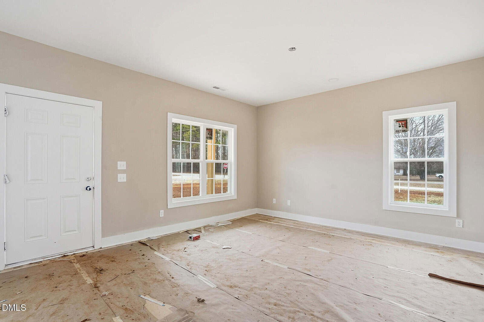 An empty room with beige walls and two large windows, covered with protective paper on the floor. A white door is on the left wall, and some tools and debris are scattered on the floor.