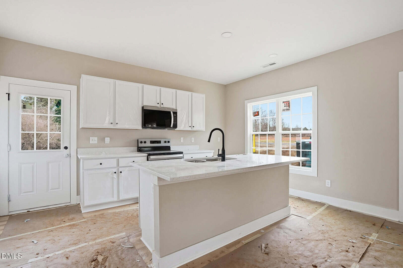 Modern, unfurnished kitchen with white cabinets, a center island with sink, stainless steel appliances, and large window letting in natural light. The floor appears unfinished and the walls are painted beige.