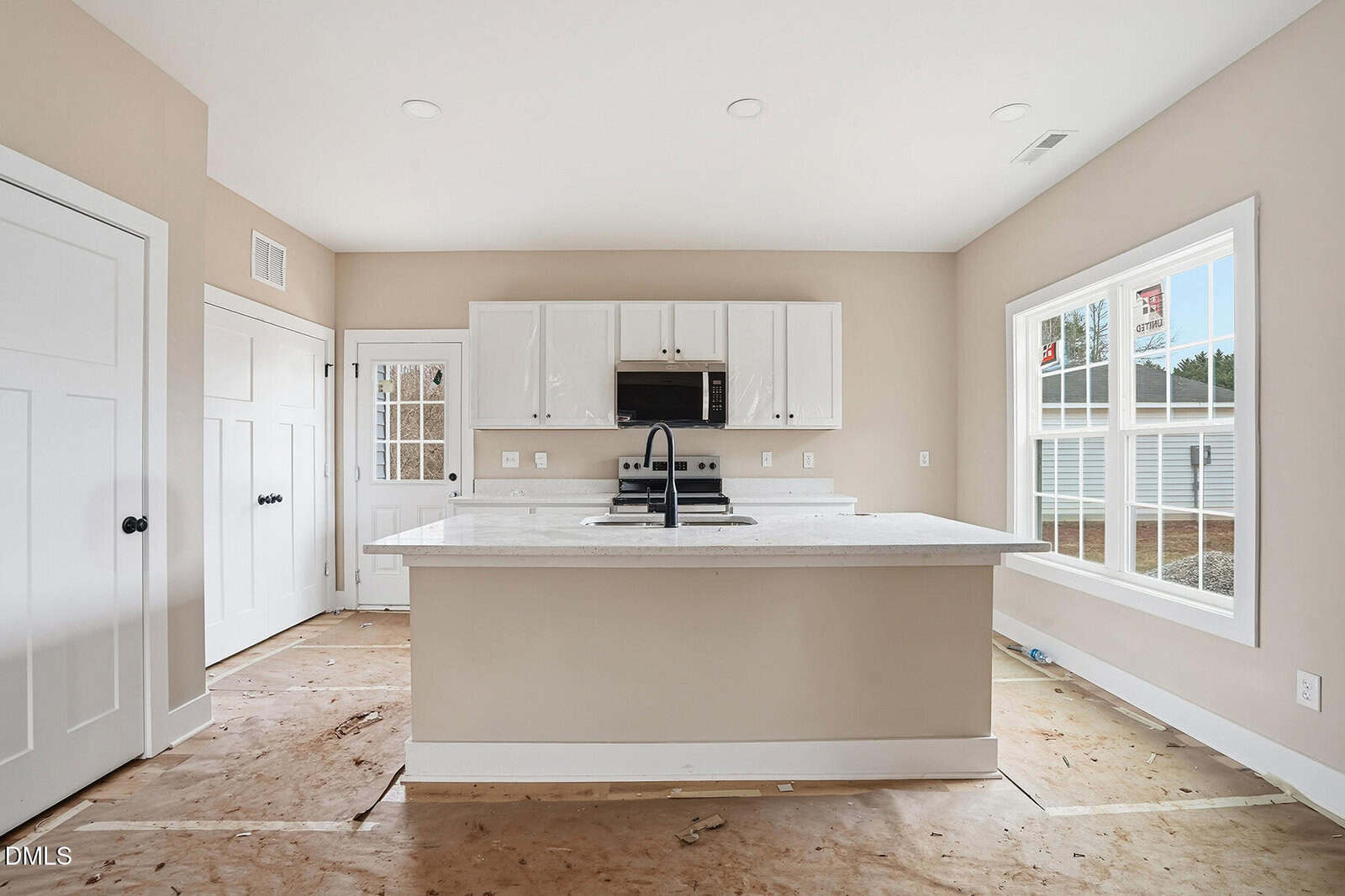 A bright, modern kitchen with white cabinets, a large island with a sink, a microwave, and an electric stovetop. The room has large windows, beige walls, and unfinished floors.