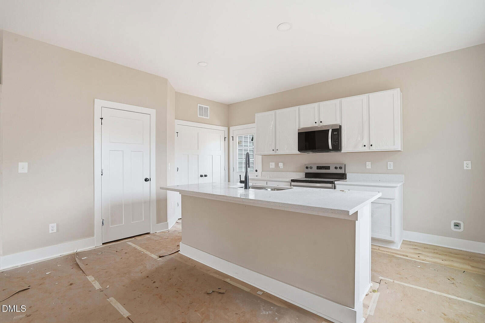 Unfinished kitchen with white cabinets, an island, stainless steel appliances, and light beige walls. The floor is covered with cardboard for protection during construction or renovation.