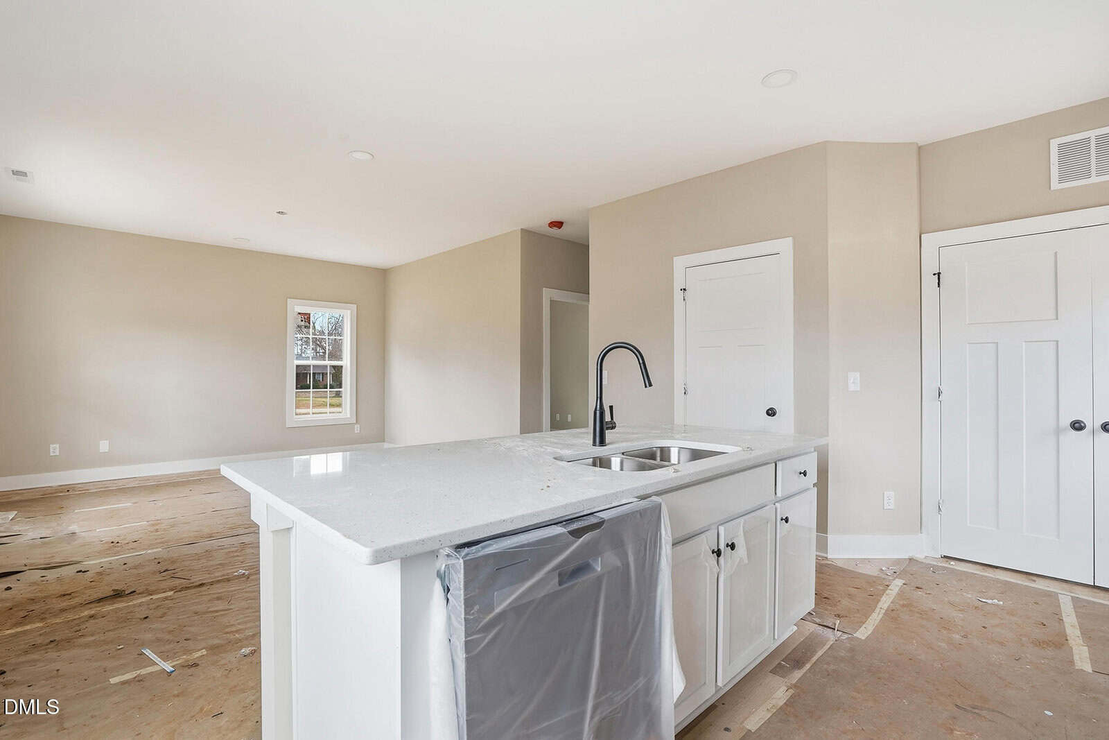 A new, unfinished kitchen with a white island, stainless steel sink, black faucet, and white cabinets. Floors are bare with construction paper covering, and walls are painted light beige.