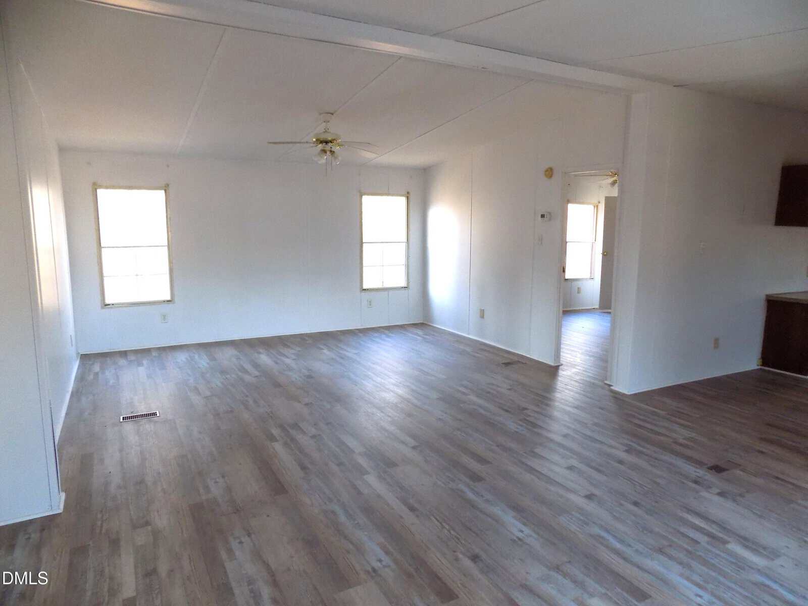 Empty room with light wood flooring, white walls, two windows, a ceiling fan with lights, and a visible doorway leading to another space; part of a kitchen counter is seen on the right.