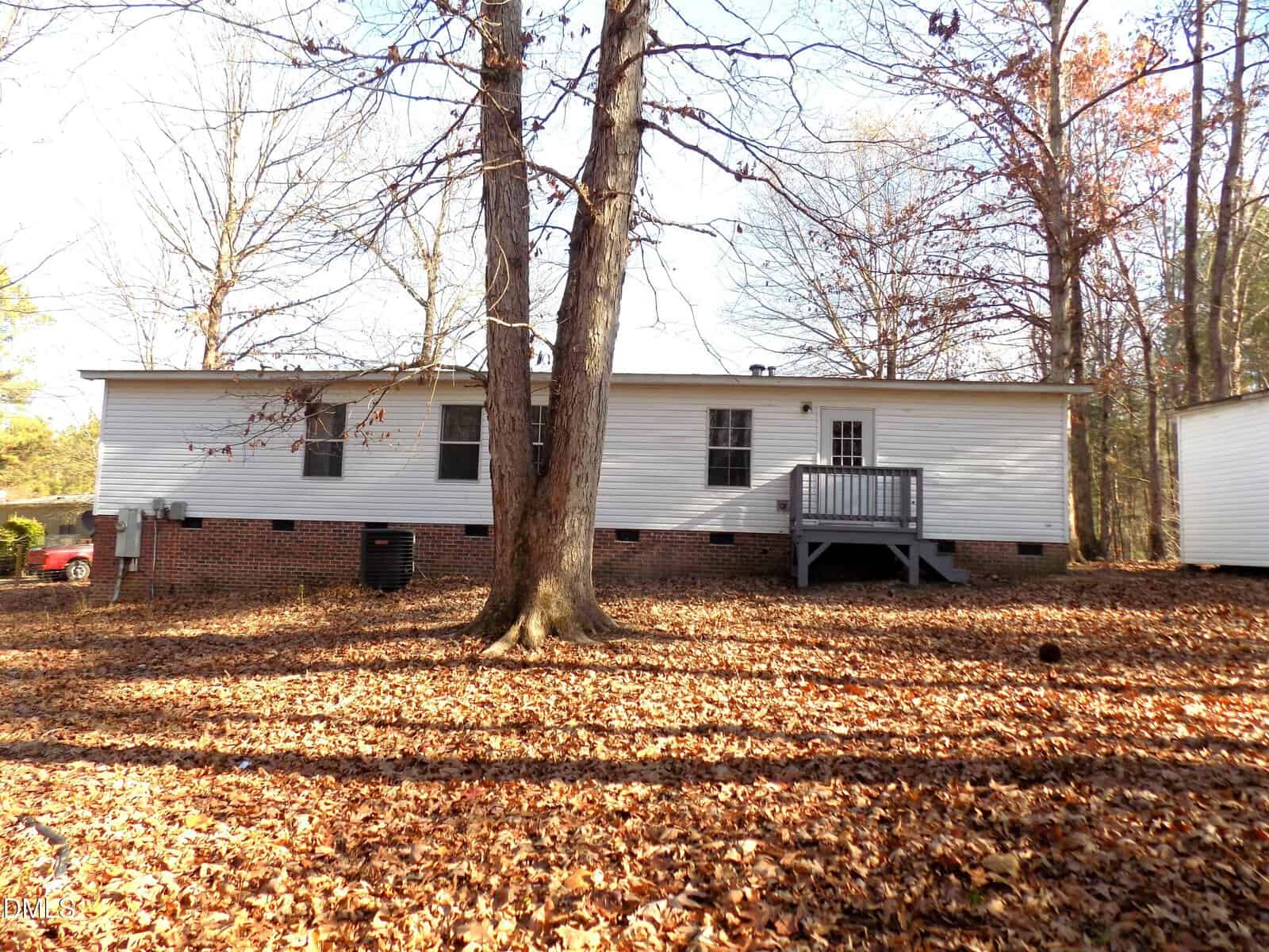 A single-story house with white siding and a brick foundation sits among tall trees. A small porch with steps leads to a back door. The ground is covered with dry fallen leaves.