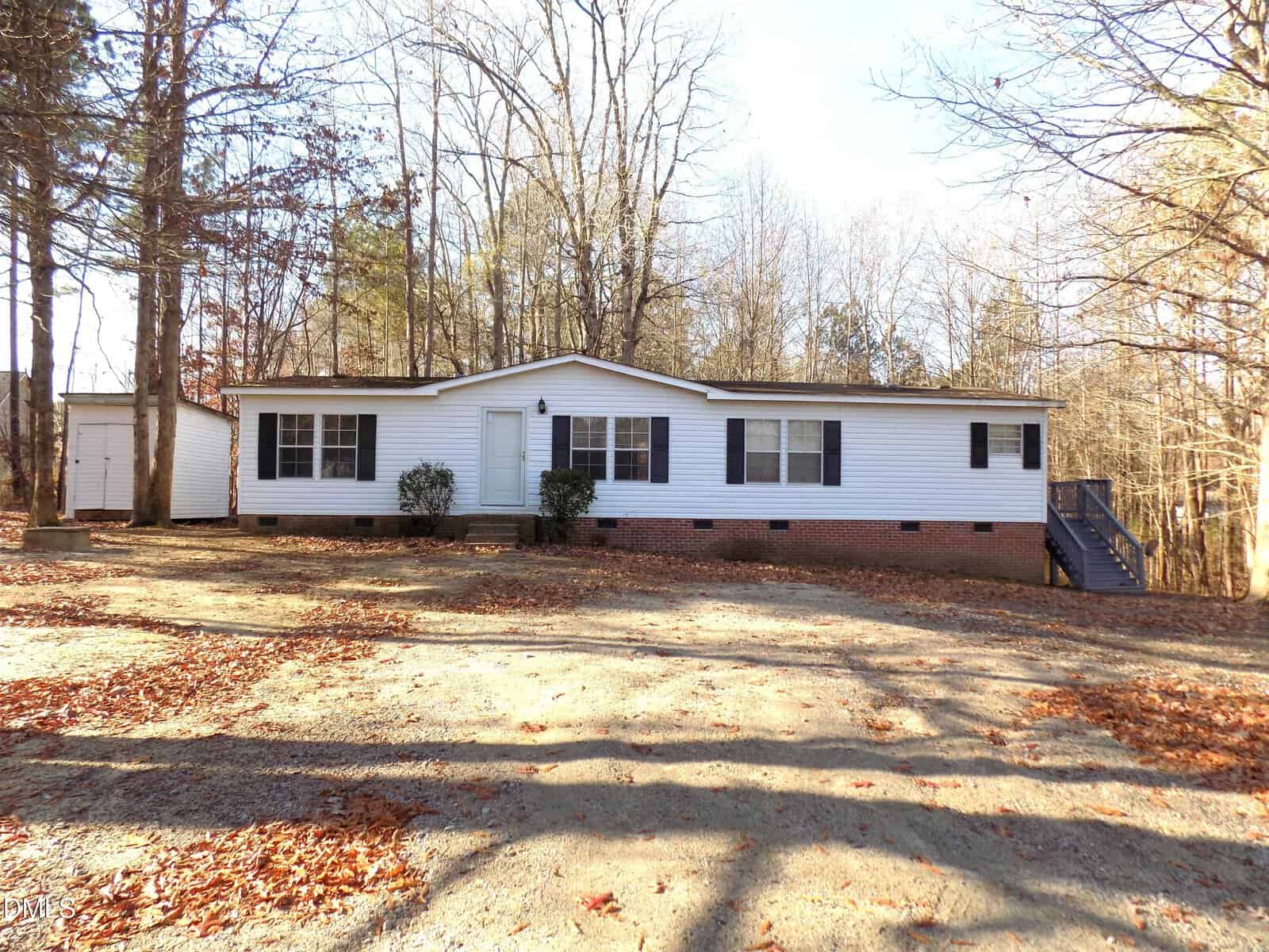 Charming white single-story manufactured home at 130 Buck Ridge Road, Louisburg, featuring a front door centered between two windows, a cozy porch with steps, and a detached garage nestled in a wooded area with leaf-covered ground and bare trees.