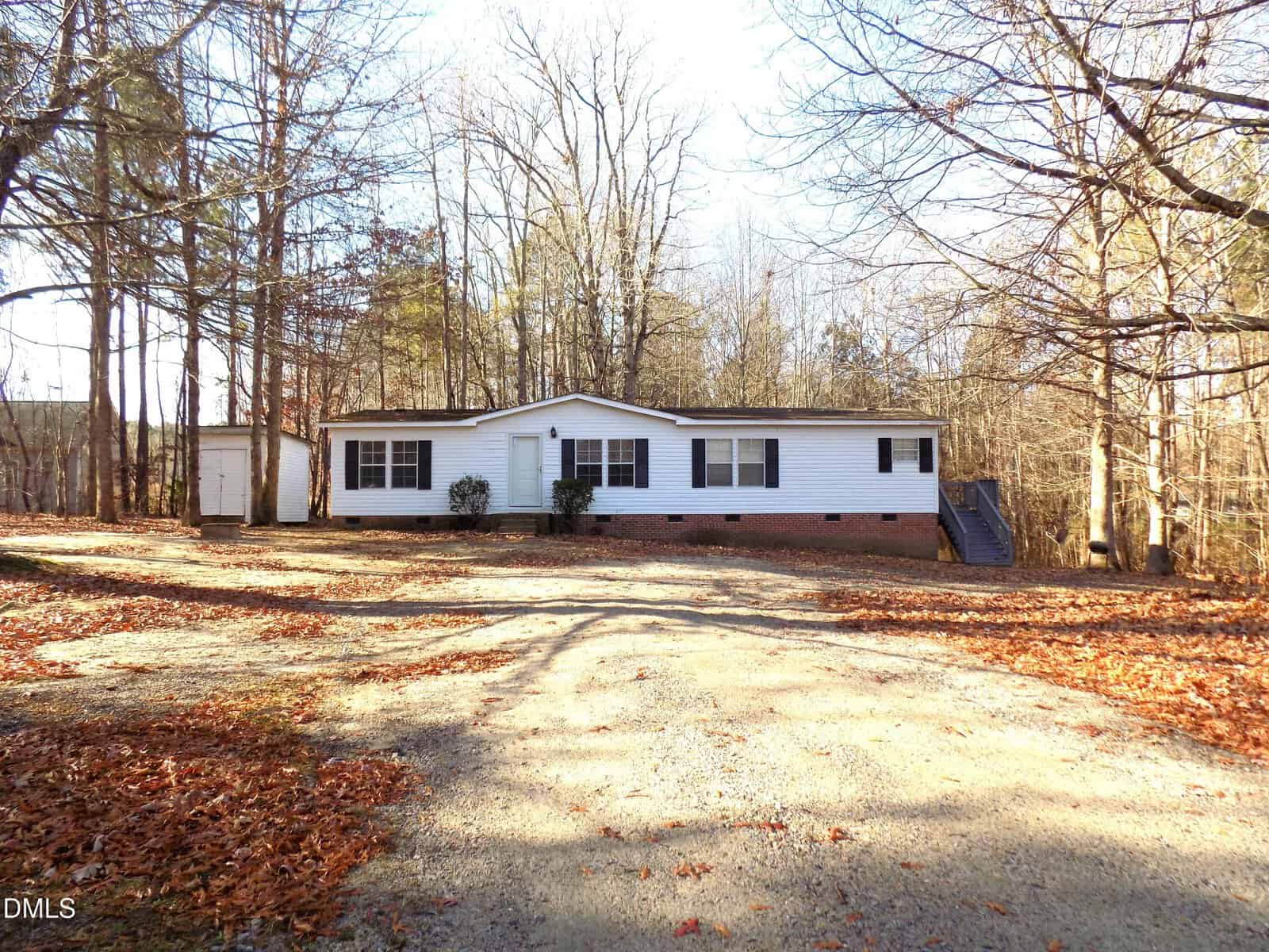 A white, single-story manufactured home with a small shed sits on a wooded lot. A gravel driveway leads to the entrance, and bare trees surround the property. Steps are visible on the right side of the house.