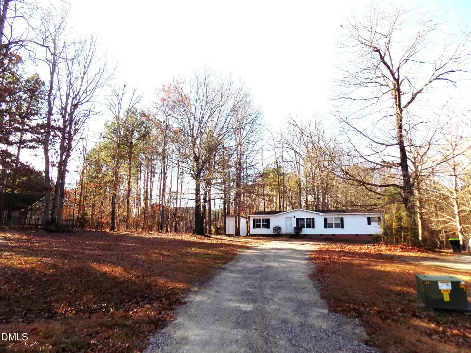 A single-story white house with black shutters sits at the end of a gravel driveway, surrounded by leafless trees and a lawn covered with scattered brown leaves under a clear sky.
