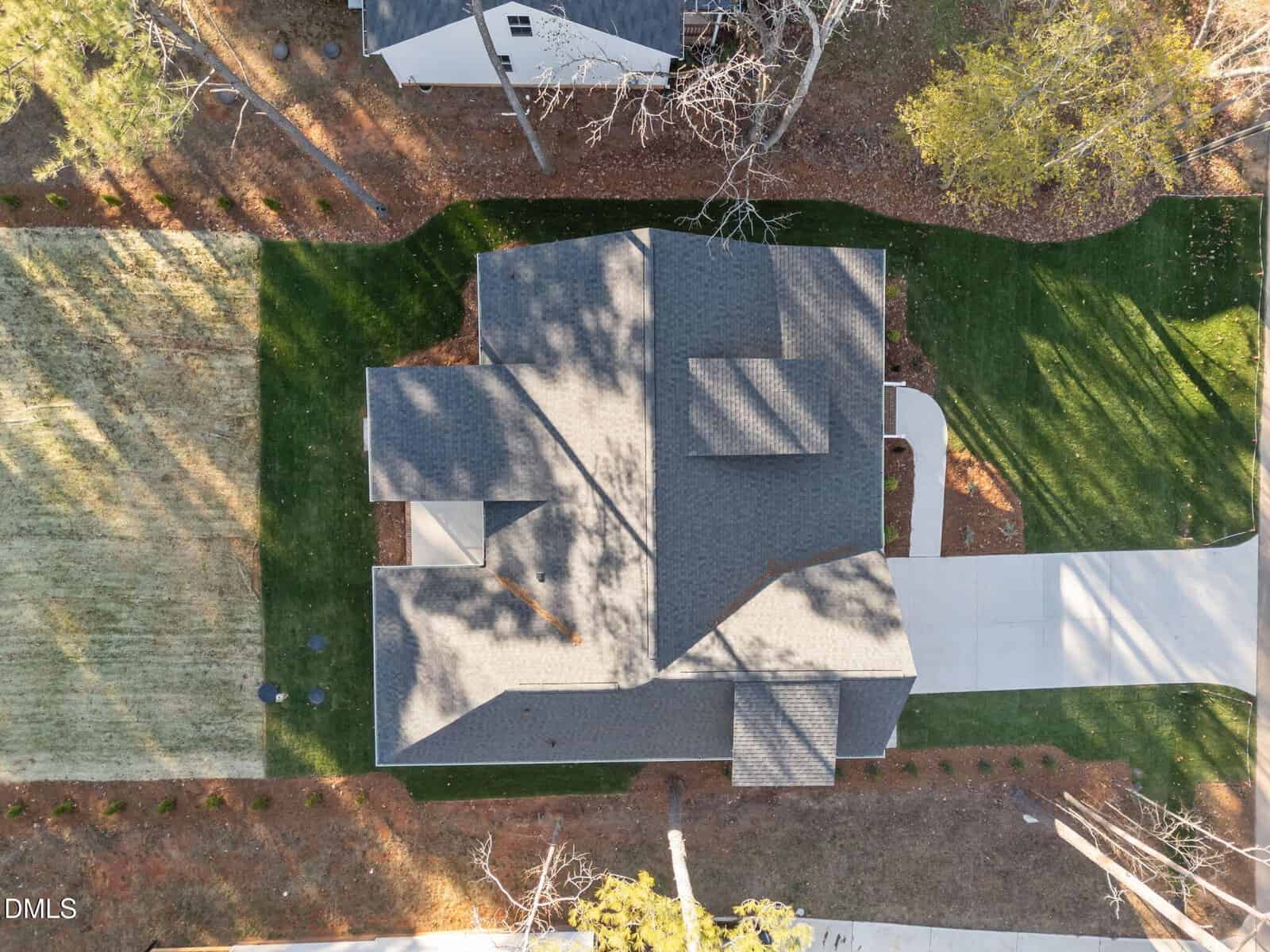 Aerial view of a gray-roofed house with a driveway, green lawns, and surrounding trees casting shadows. The house sits centrally with neatly landscaped areas and another house visible at the top of the image.