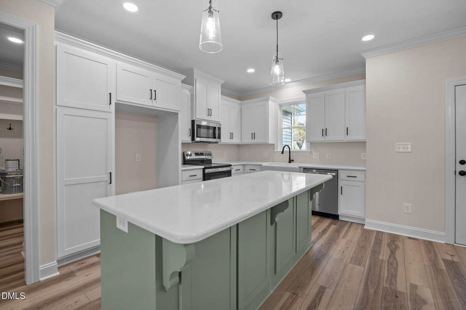Bright, modern kitchen with white cabinets, stainless steel appliances, a large white marble island with green base, pendant lights, wood flooring, and natural light from a window above the sink.