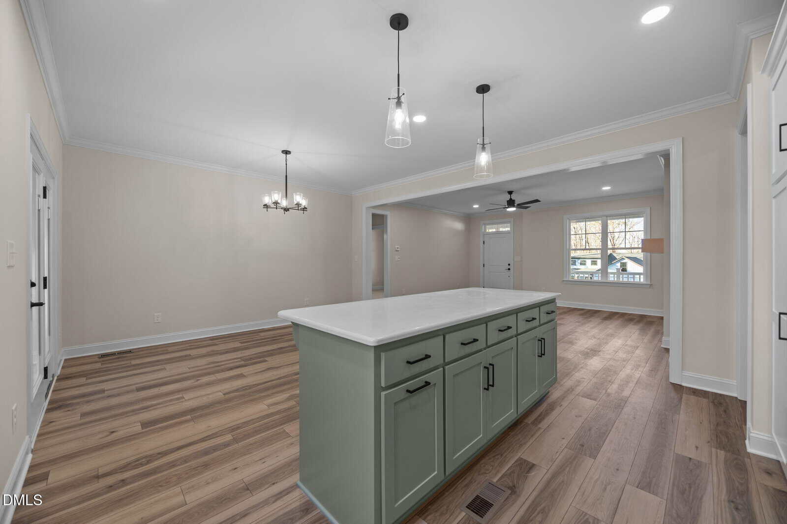 Open kitchen and dining area with light wood floors, an island with green cabinets and white countertop, pendant lights above, neutral walls, and a view into the living room with large windows.