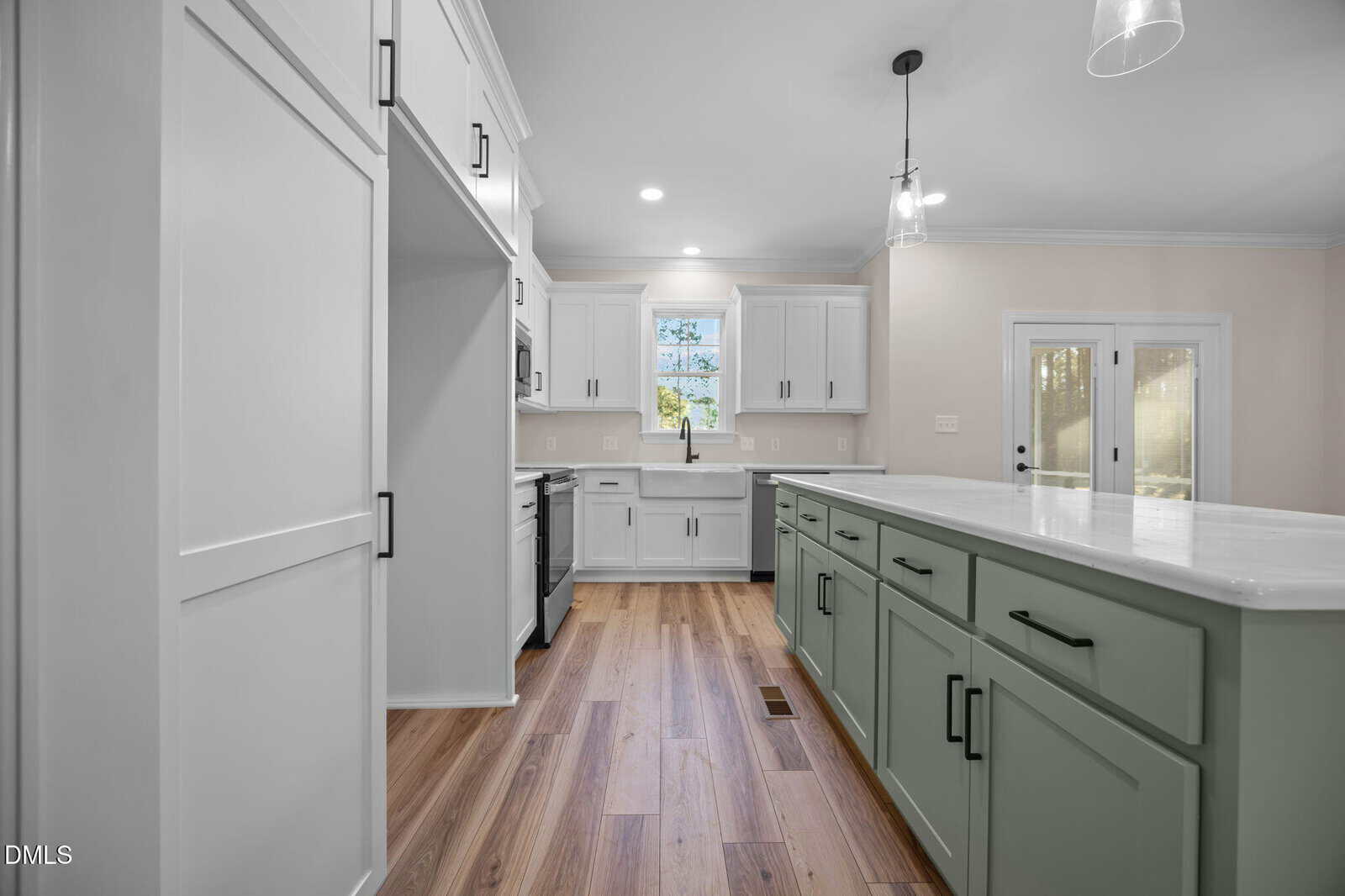 Modern kitchen with white upper cabinets, light green island with drawers, wood flooring, farmhouse sink under a window, pendant lights, and double glass doors leading outside.