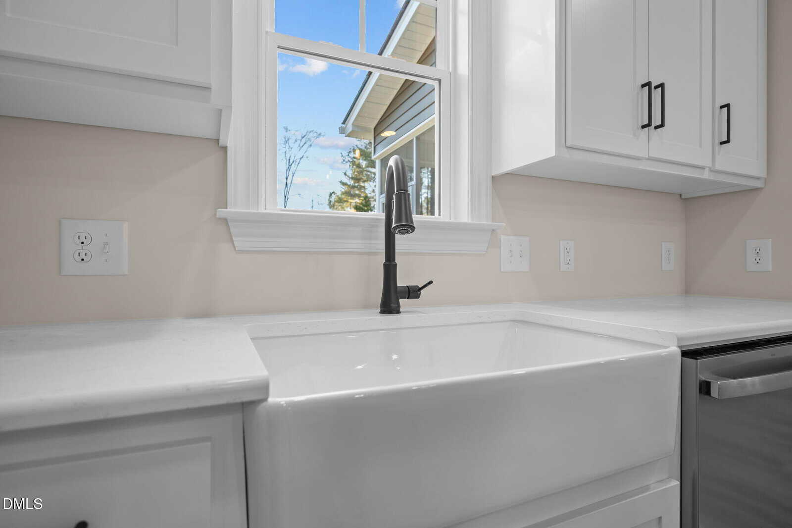 Close-up of a modern kitchen sink with a black faucet, white cabinets, and white countertops beneath a window showing blue sky and trees. Multiple electrical outlets are visible on the backsplash.