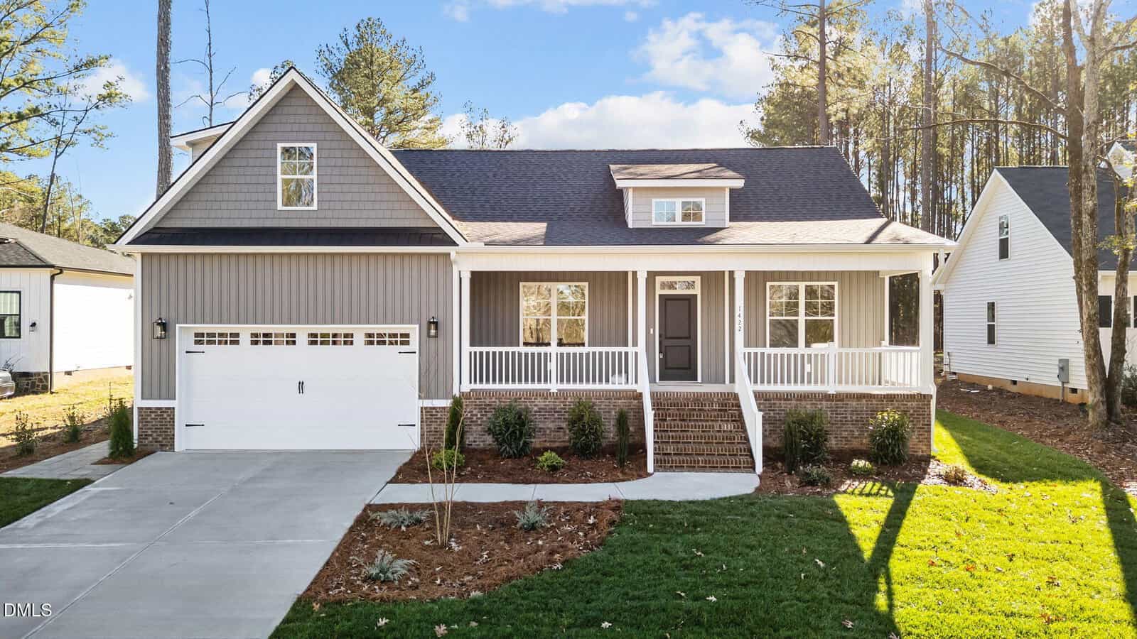 A modern two-story house with gray siding, white trim, a covered front porch, and attached two-car garage. The yard is landscaped with grass, shrubs, and a concrete driveway. Tall trees are visible in the background.