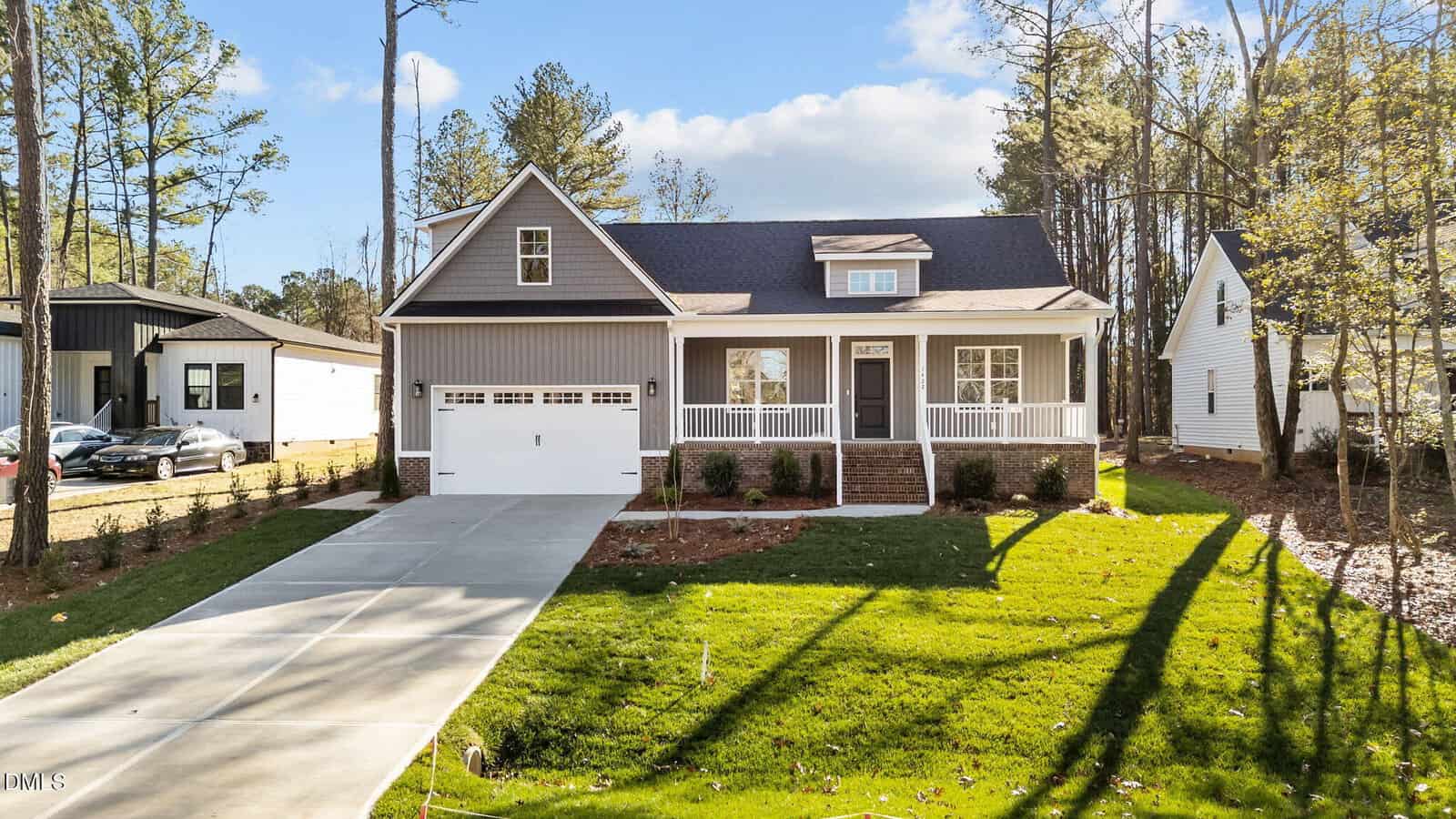 A modern single-story house with a gray exterior, white trim, a covered front porch, and an attached two-car garage. The home has a well-kept lawn, a concrete driveway, and is surrounded by tall trees.