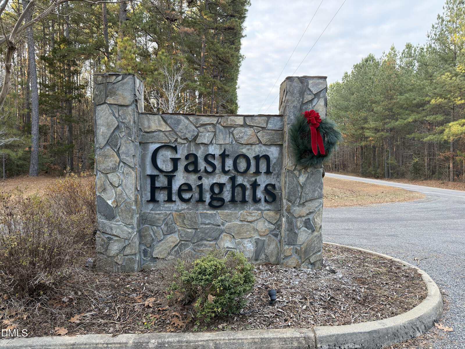 Stone entrance sign reading “Gaston Heights” with a seasonal wreath, surrounded by trees, bushes, and a curved road on a cloudy day at 0 Woodland Rd., Littleton.