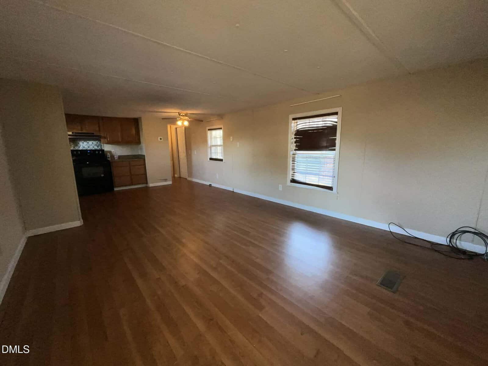 Spacious empty living area with wood flooring, beige walls, and a window with blinds. In the background, there is an open kitchen with wooden cabinets and black appliances. A bundle of cables rests on the floor near the window.