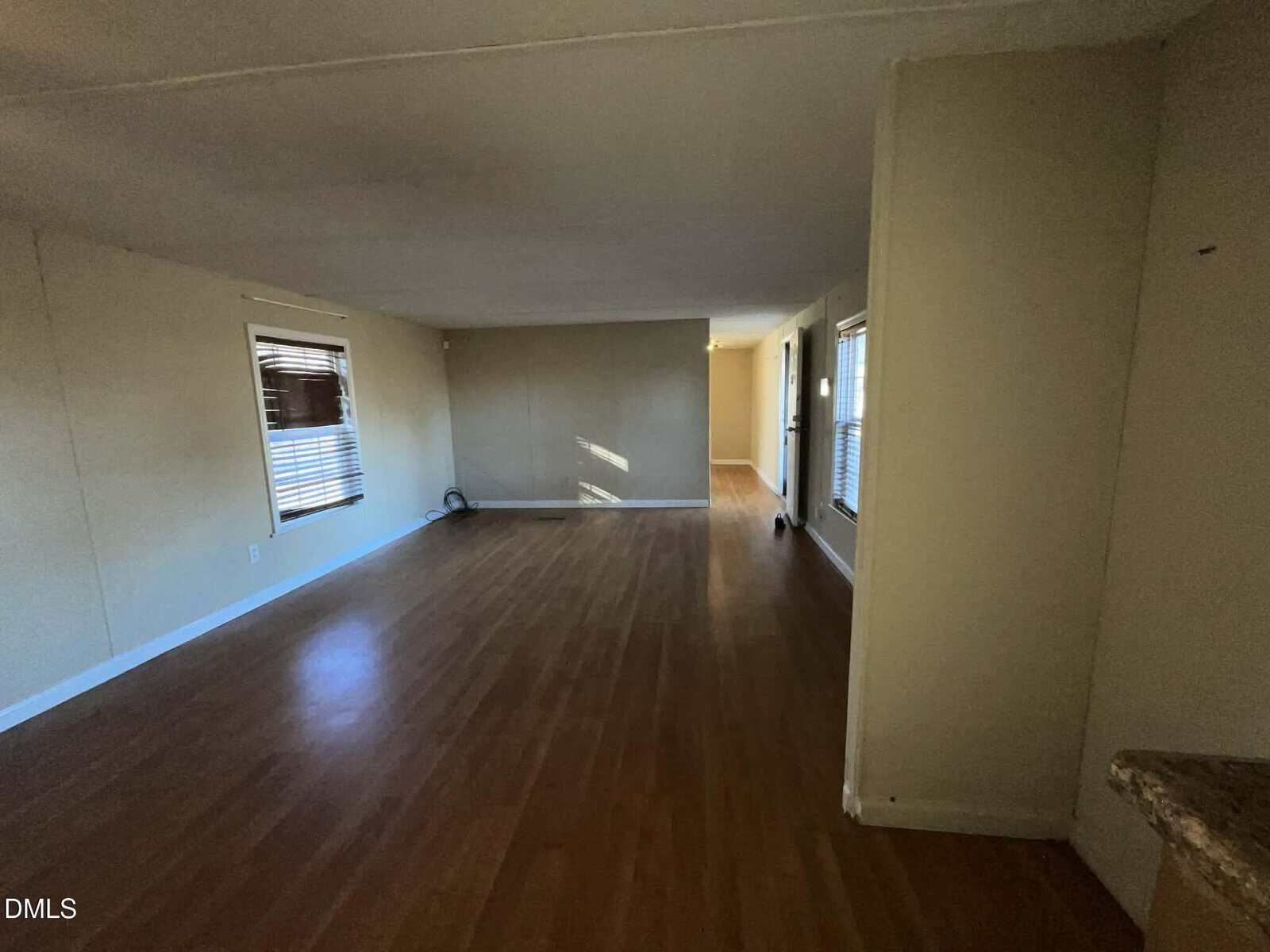 Spacious, empty room with light-colored walls and wood laminate flooring, featuring multiple windows with blinds allowing natural light and an open layout extending toward another room.