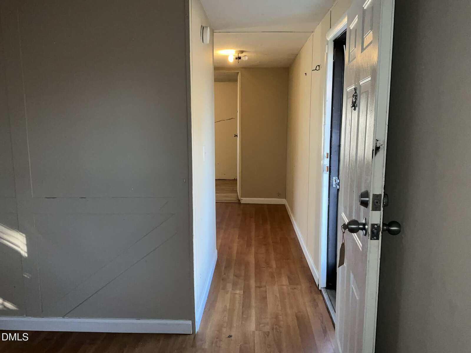 A hallway with light wood flooring, beige walls, an open white front door on the right, and a lit ceiling fixture at the end, leading to closed doors in the background.