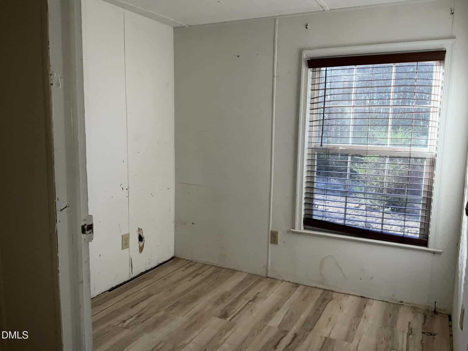 Small empty room with white walls, light wood flooring, a large window with blinds, and some marks or scuffs on the walls. Natural light enters through the window.