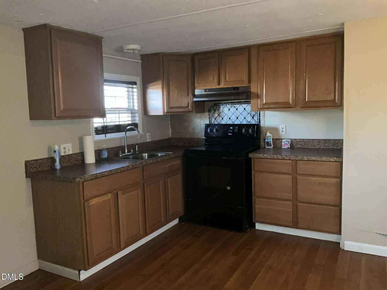 A small kitchen with wooden cabinets, a black electric stove, stainless steel sink under a window with blinds, brown countertops, and a wood floor. Cleaning supplies and a paper towel roll are on the counter.