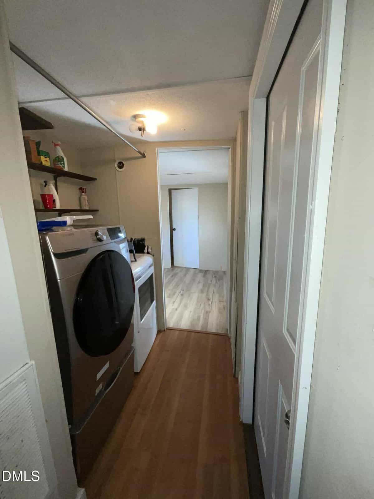 A narrow laundry area with a front-loading washing machine and dryer on the left, open shelves with cleaning supplies above, and a hallway leading to a bright room with wood flooring.