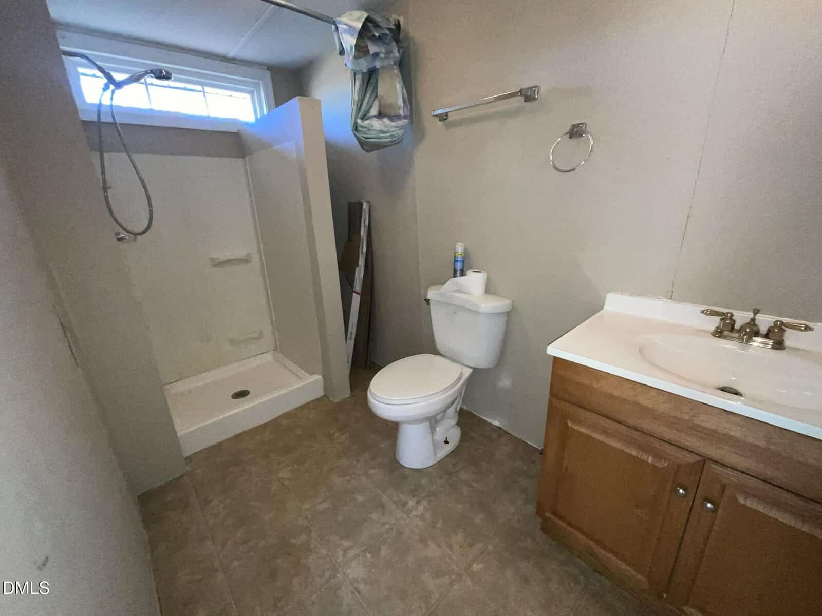 A bathroom with a walk-in shower, toilet, and wooden vanity with sink. Towels hang above the shower, and toiletries are on the toilet tank. The floor is tile, and the walls are light-colored.
