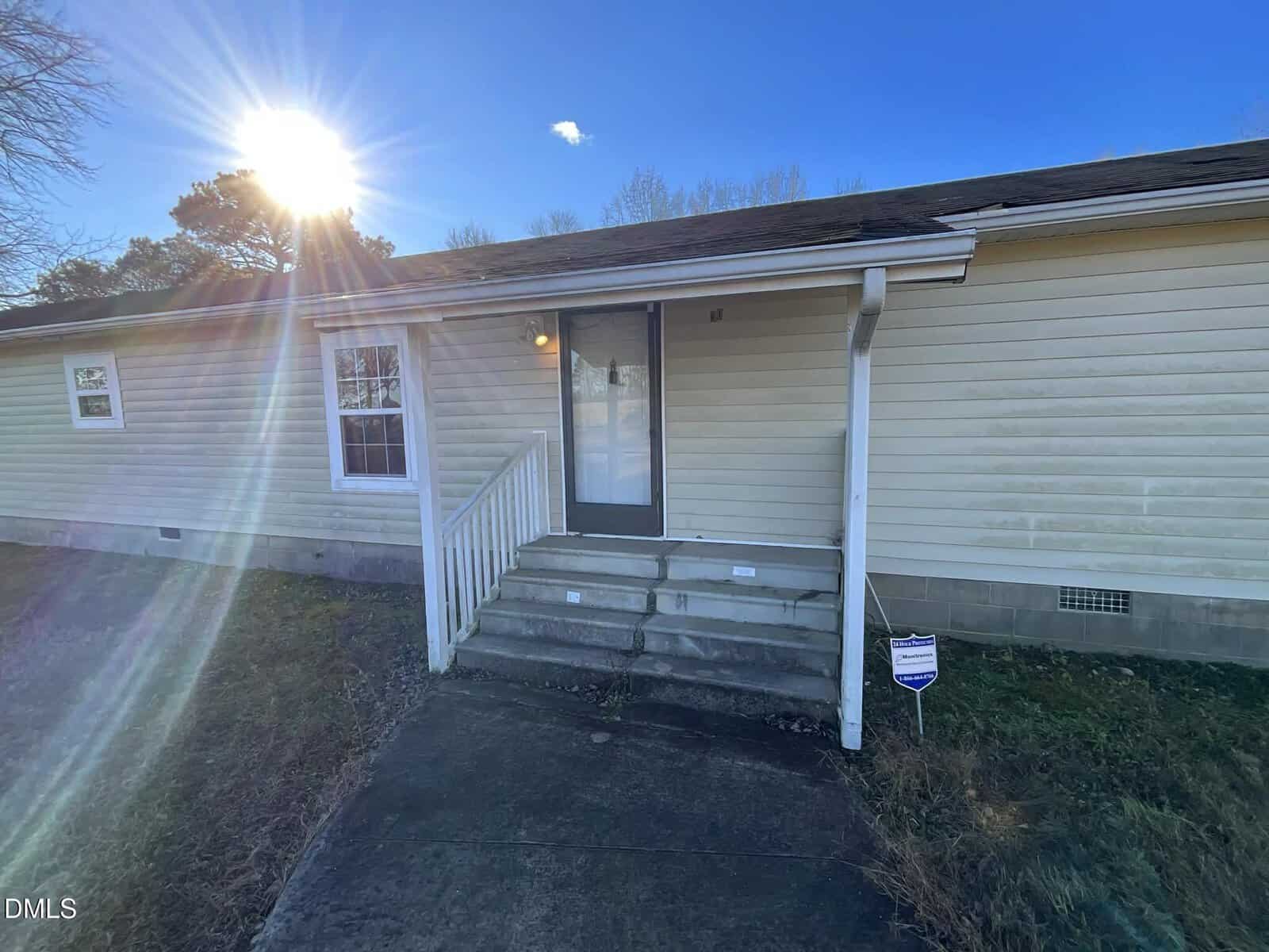 A beige house with a small front porch and concrete steps leading to a glass door. The sun shines brightly in the blue sky, casting lens flare across the yard. A security sign is visible near the steps.