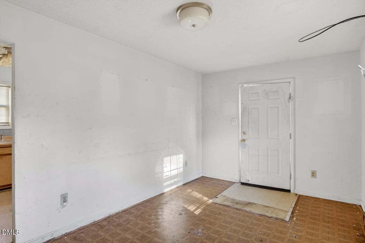 An empty room with white walls, a ceiling light, a door with a window, and a worn-out floor with visible stains and patches. Part of a kitchen with wooden cabinets is visible through an open doorway on the left.