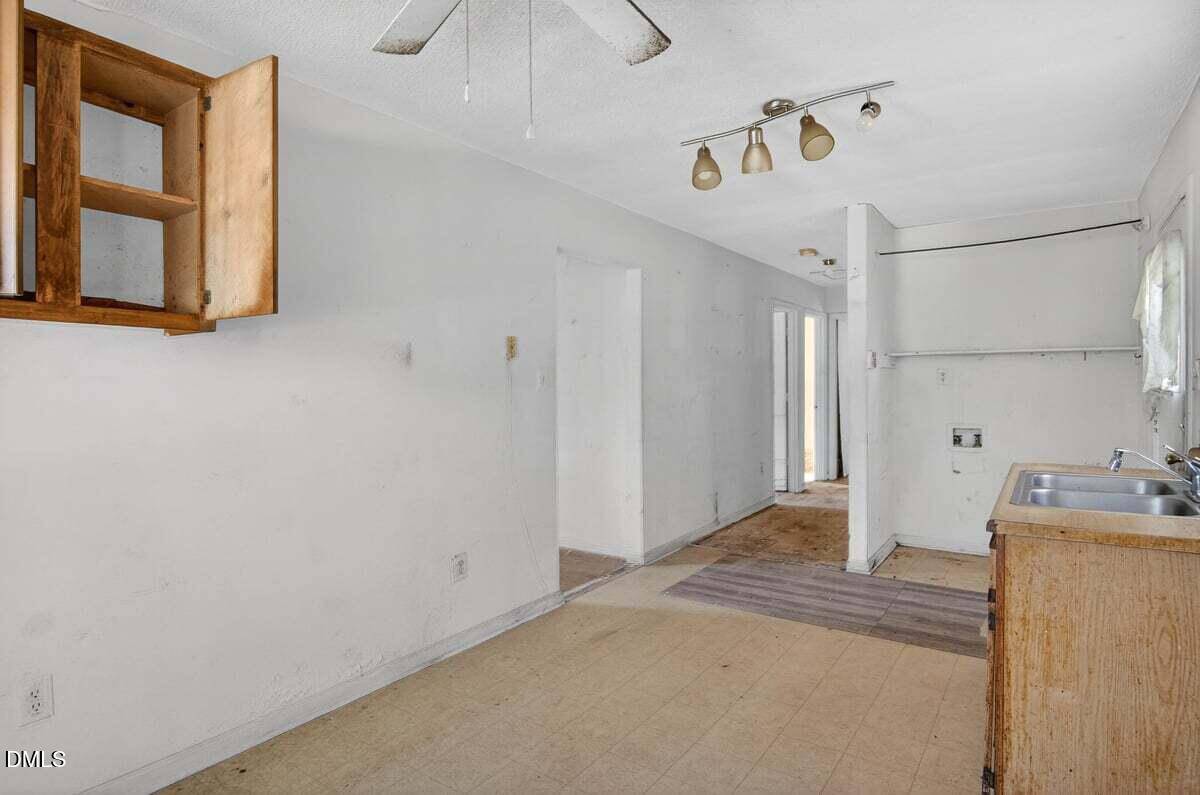 A worn, empty kitchen with light-colored walls, an open wooden cabinet, a double sink, basic counters, visible plumbing connections, and a ceiling fan. The room leads to a hallway with multiple doorways.