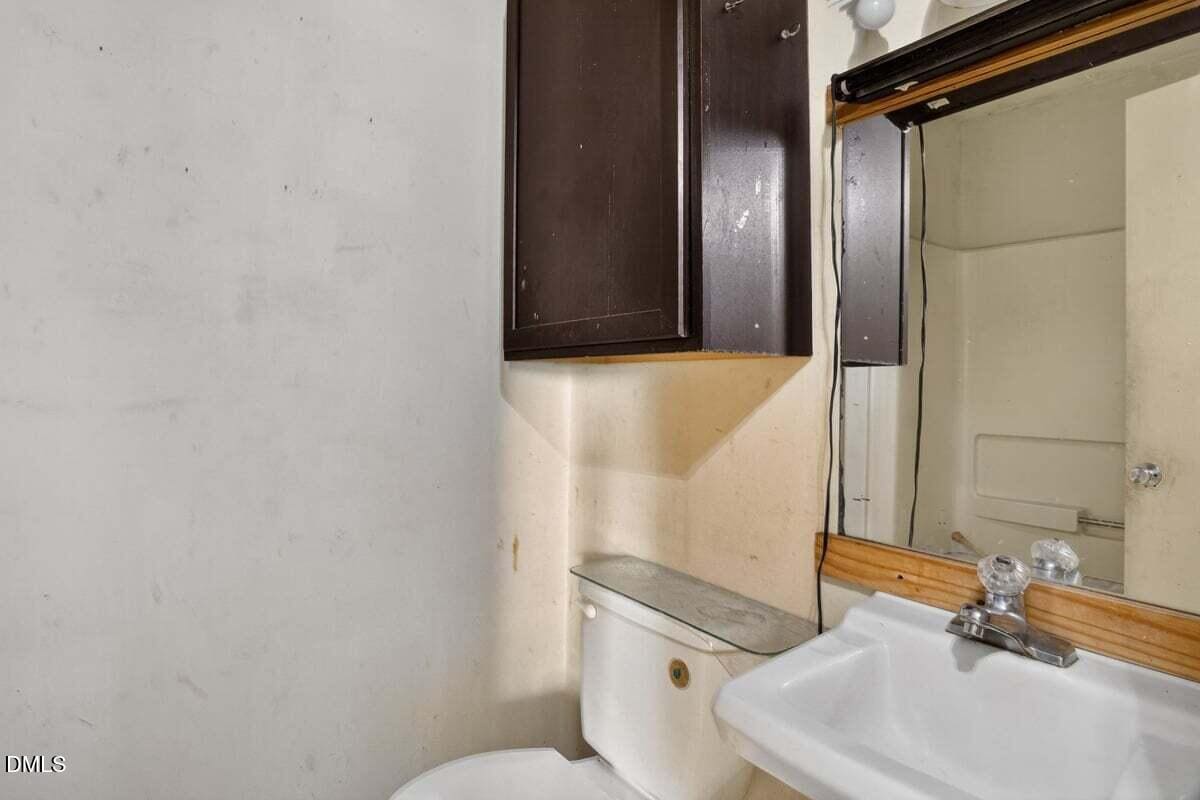 Small bathroom with a white sink, faucet, and mirror above. A toilet is beneath a dark wall cabinet. The walls appear worn, and part of a bathtub is visible in the mirror’s reflection.