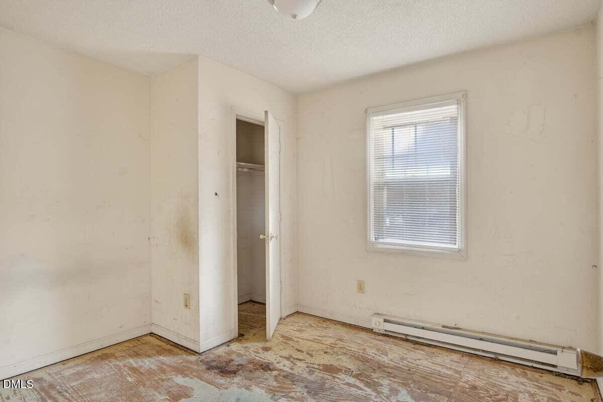 Small empty room with stained walls, an open closet, dirty floor with missing carpet, a single window with blinds, and a baseboard heater below the window. The space appears to be in need of renovation.